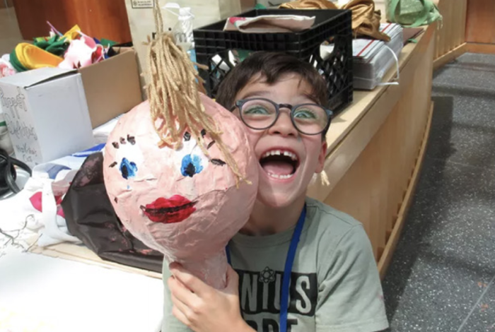 Smiling boy with glasses holding up the paper mache round head he created in a MAM puppetry course.
